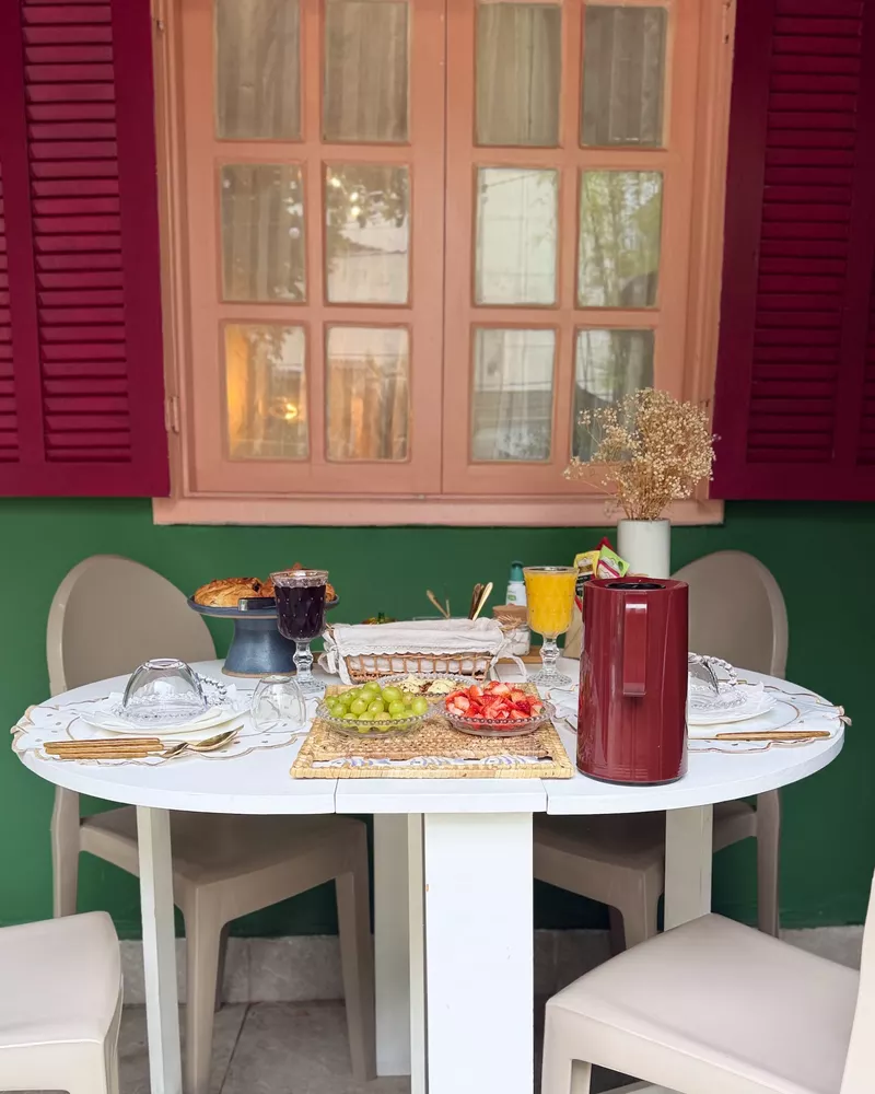Mesa branca com cadeiras diante de uma janela rosa com venezianas vinho.